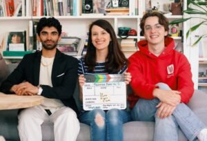 a photo showing three people sitting on a couch in front of a bookcase: a man with a beard wearing a black blazer, a woman with long dark hair and a striped shirt holding a clapper and a man with glasses wearing a red sweatshirt